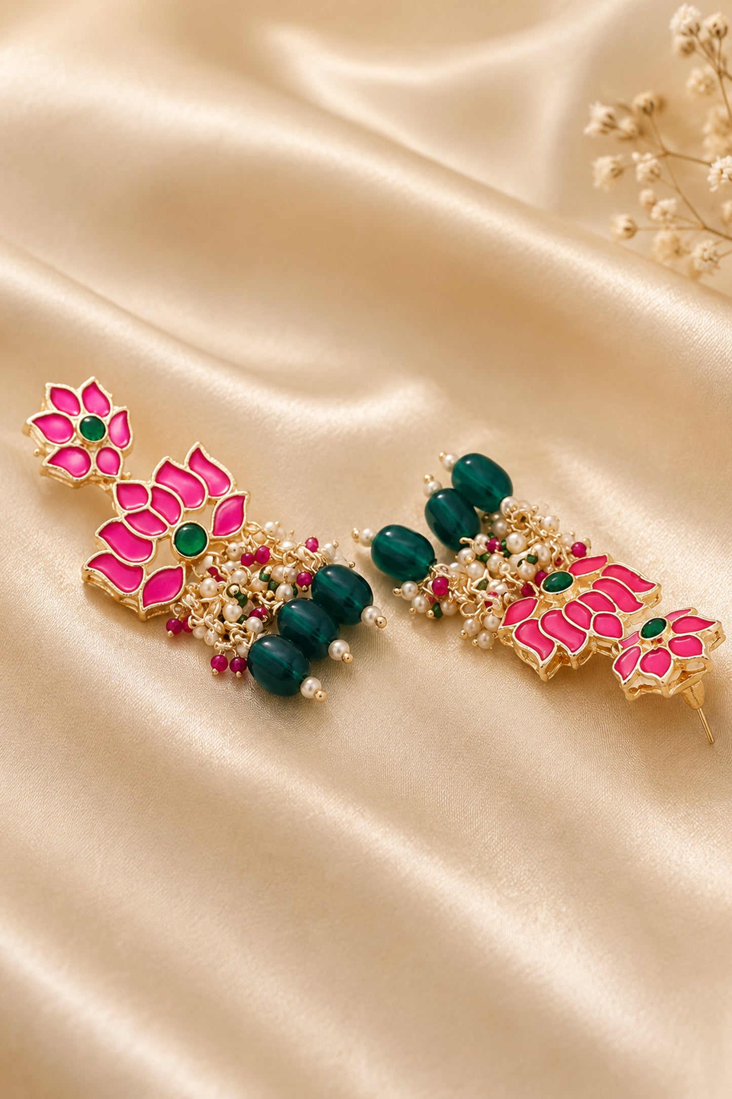 A pair of ornate gold-toned statement earrings with bright pink lotus flower motifs, green stones, and dark green bead accents, arranged on a draped champagne satin fabric.