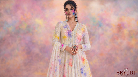 Woman in a white embroidered anarkali dress with colorful Holi powder on her face and outfit, standing against a blurred background of vibrant color clouds.
