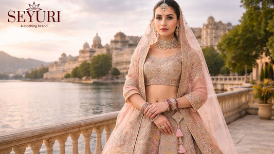 Elegant South Asian bride in a pastel peach embroidered lehenga with gold jewelry, standing by a lakeside terrace with palace architecture at sunset.