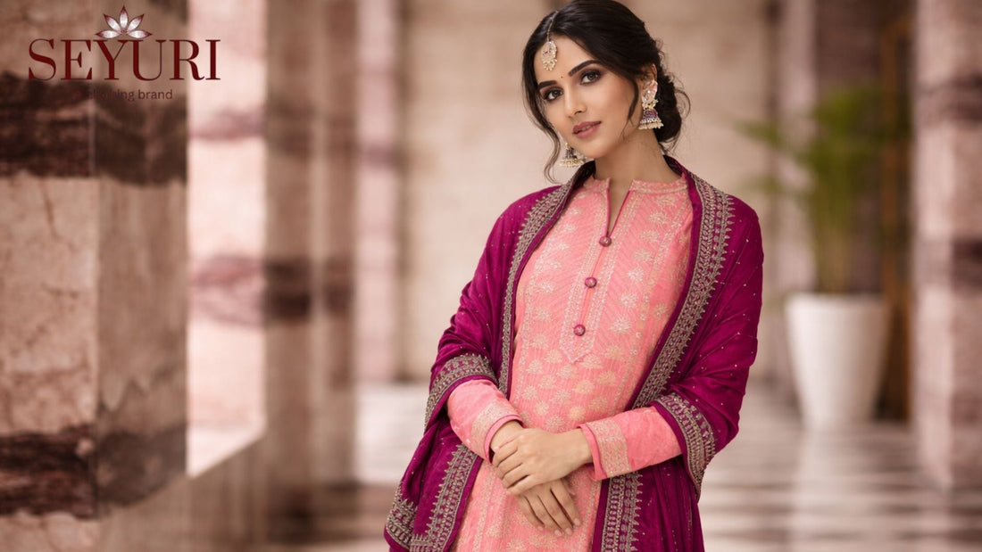 Woman wearing a pastel pink embroidered salwar kameez with a deep magenta dupatta, styled with traditional jewelry, standing in a softly blurred marble corridor, highlighting the outfit’s intricate detailing and graceful silhouette.