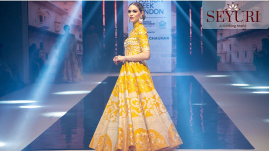 A woman wearing a vibrant yellow and pink floral lehenga, smiling in front of a traditional heritage palace and garden.