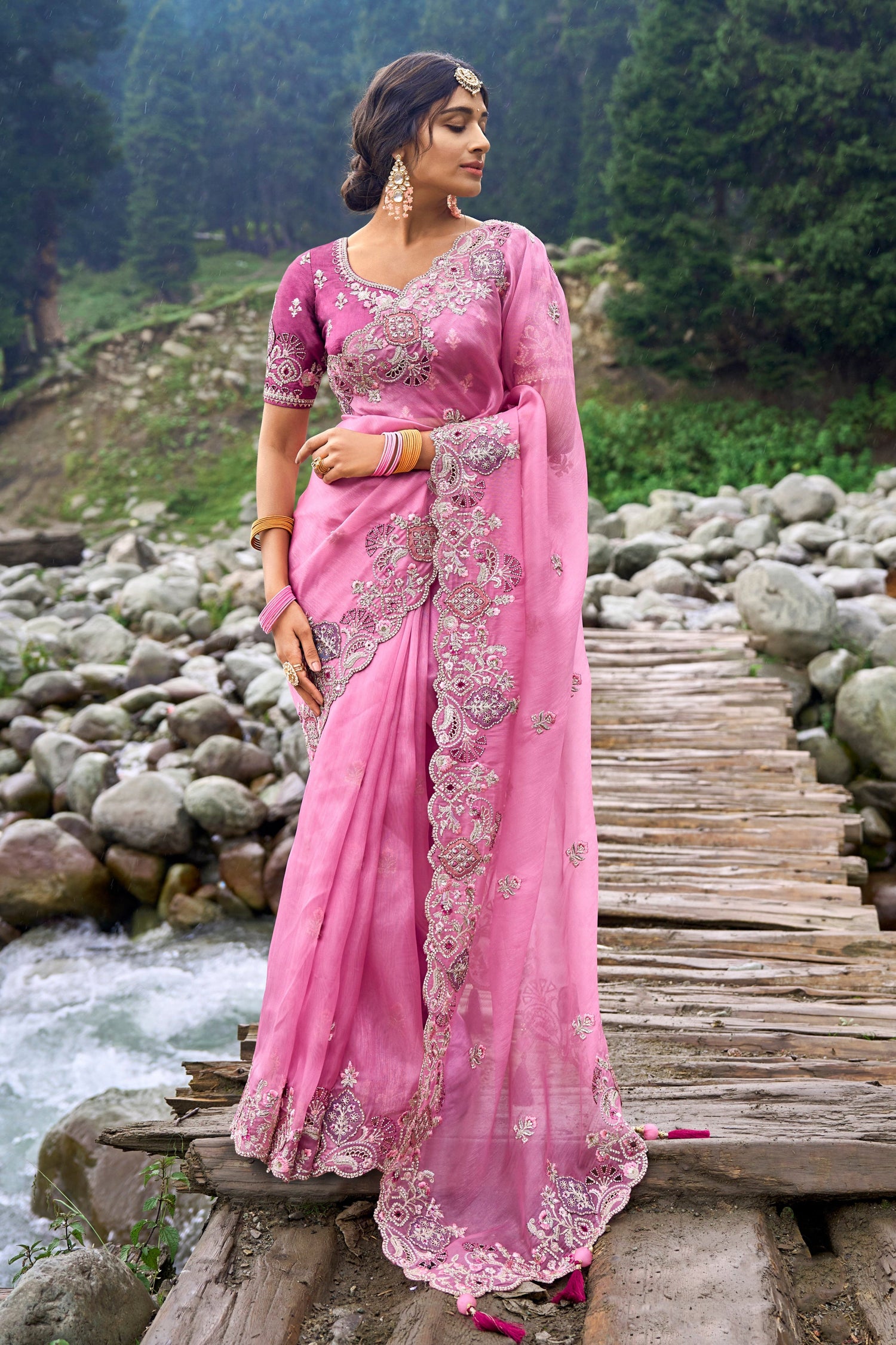 Woman in a pink saree standing on a wooden bridge over a stream with trees in the background