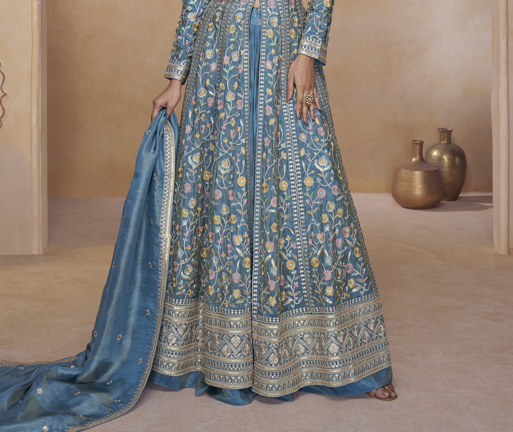 Blue Embroidered Floor-Length Gown with Detailed Floral Work, Long Sleeves, and Matching Dupatta Worn by a Woman at a Royal Backdrop.
