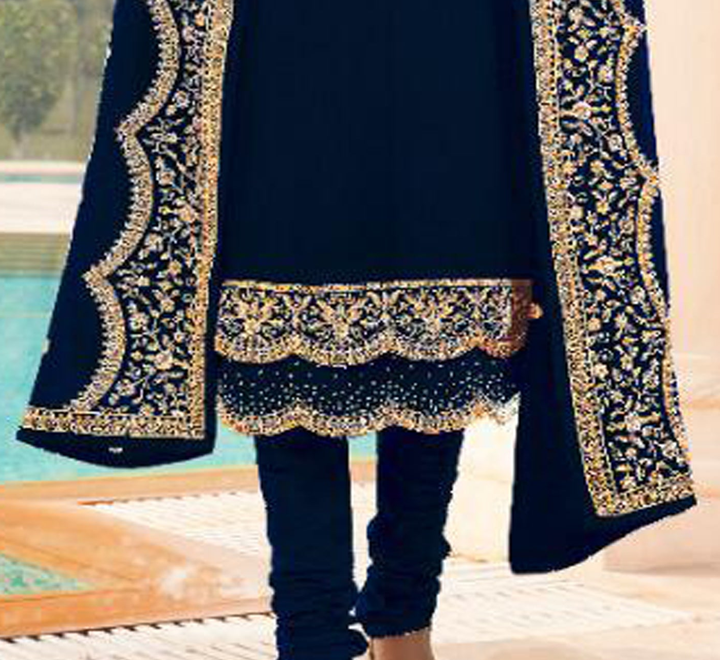 A person wearing a black traditional outfit with ornate gold embroidery and beaded scalloped borders, standing near a poolside setting.