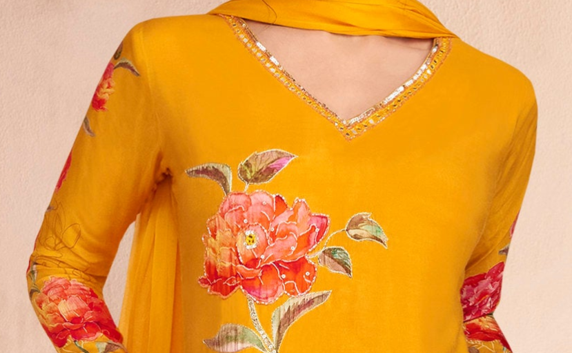 A close-up of a mustard yellow traditional outfit with a large pink and red floral embroidery on the front, featuring a V-neckline with gold trim and matching floral embroidery on the sleeves.