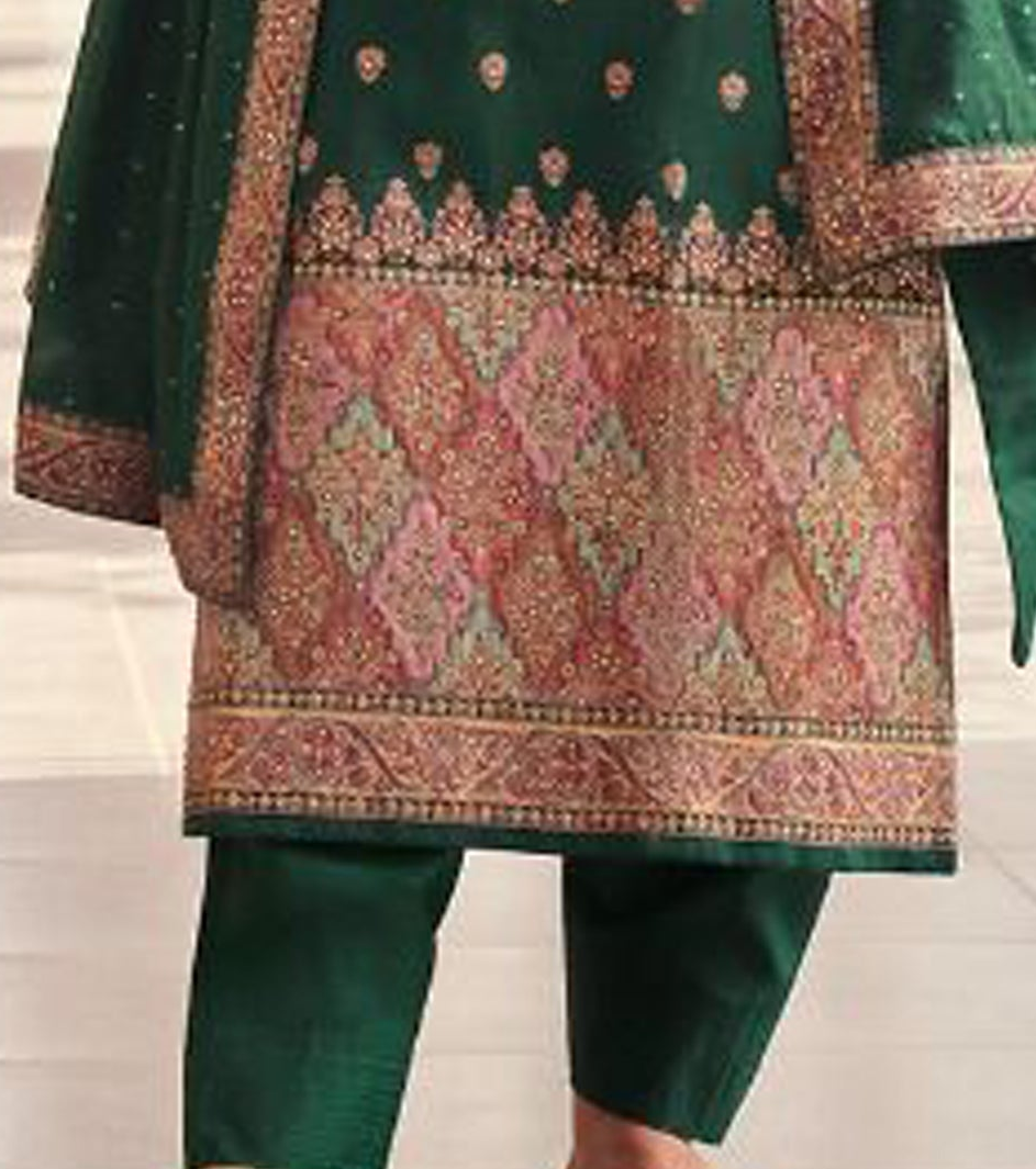 Woman in a green traditional outfit with intricate patterns standing indoors.