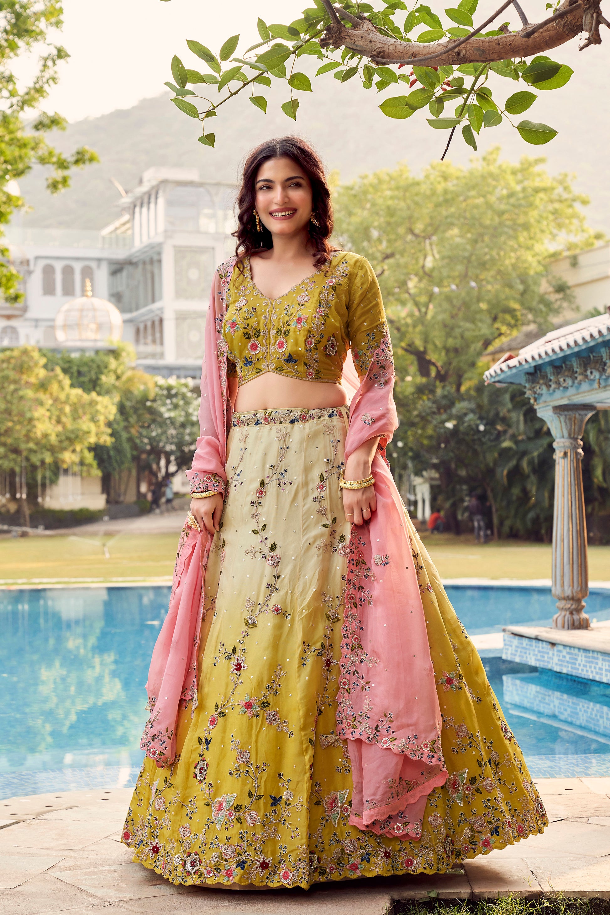 Woman Wearing a Vibrant Yellow Floral Embroidered Lehenga With a Pink Dupatta, Posing By A Serene Poolside.