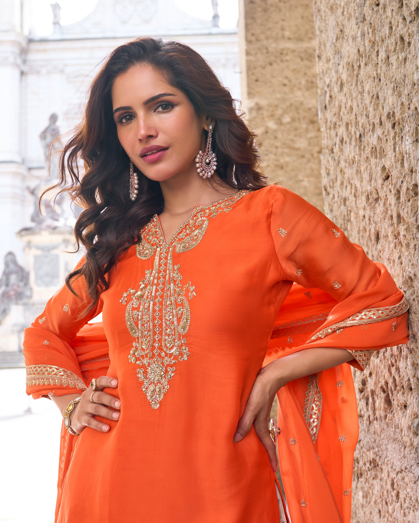Woman wearing an orange traditional outfit with intricate designs, standing against a blurred background.