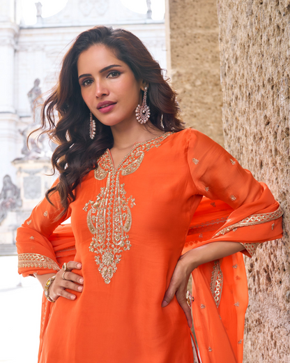 Woman wearing an orange traditional outfit with intricate designs, standing against a blurred background.