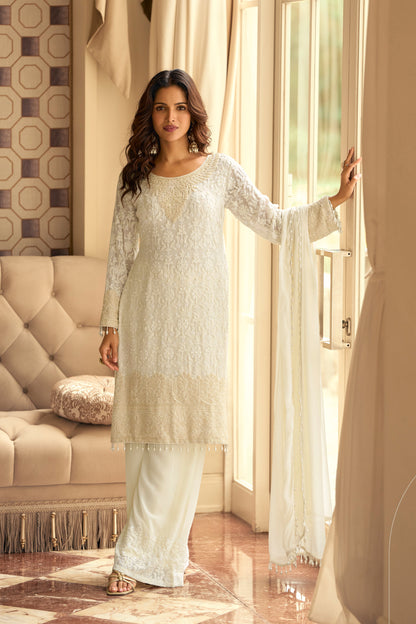 Woman in a white traditional outfit standing in a well-lit room with a couch and decorative pillows.