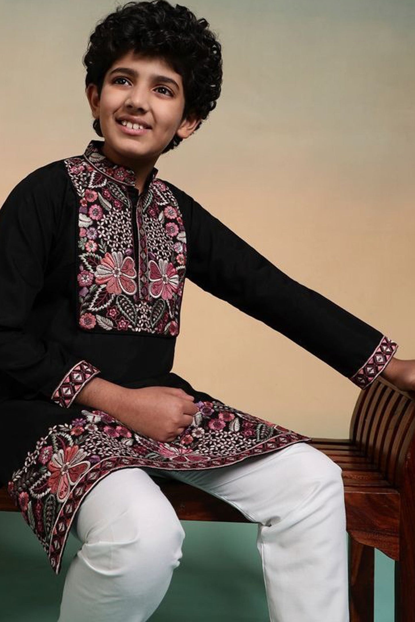 Child wearing a traditional embroidered kurta with white pants, sitting on a wooden bench.